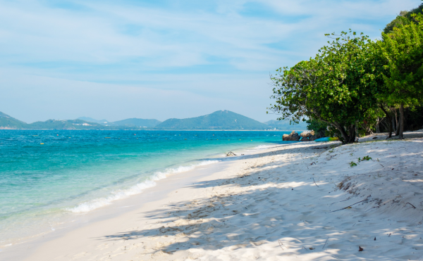 Plage en thailande avec une eau bleu, une plage de sable blanc, et des arbres, en fond des iles et montagnes lointaines